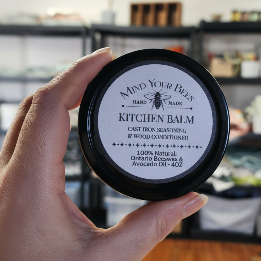Hand holding a jar of 'Mind Your Bees' Kitchen Balm with a blurred background